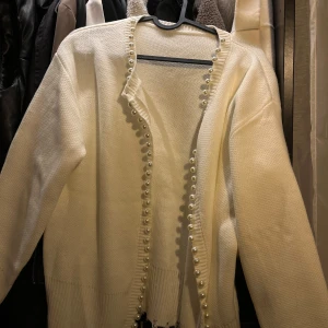 Vit kofta med pärldetaljer - Säljer en superfin vit kofta med pärldetaljer längs kanten. Den är perfekt för att ge en elegant touch till vilken outfit som helst. Koftan har långa ärmar och är i ett mjukt stickat material. Passar perfekt för både vardag och fest