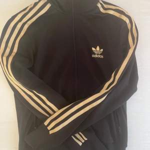 Säljer en svart Adidas jacka med de klassiska guldiga ränderna längs ärmarna. Jackan har en dragkedja framtill och Adidas-loggan på bröstet. Perfekt för en sportig look eller chill vardagsstil. Oklar storlek. Skulle säga xs. Lite sönder vid armen