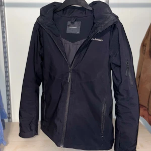 Svart vindjacka från Peak Performance - Säljer en svart vindjacka från Peak Performance. Jackan har en dragkedja framtill och är långärmad med en huva. Perfekt för blåsiga dagar och har en stilren design. Passar bra till både vardag och utomhusaktiviteter.