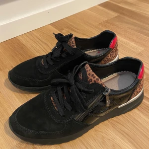 Svarta Rieker-skor, storlek 40 med leoparddetaljer - Säljer ett par svarta Rieker-skor i storlek 40. Skorna har leoparddetaljer och en röd accent vid hälen, vilket ger dem en unik och snygg design. De är utrustade med en dragkedja på sidan för enkel på- och avtagning.
