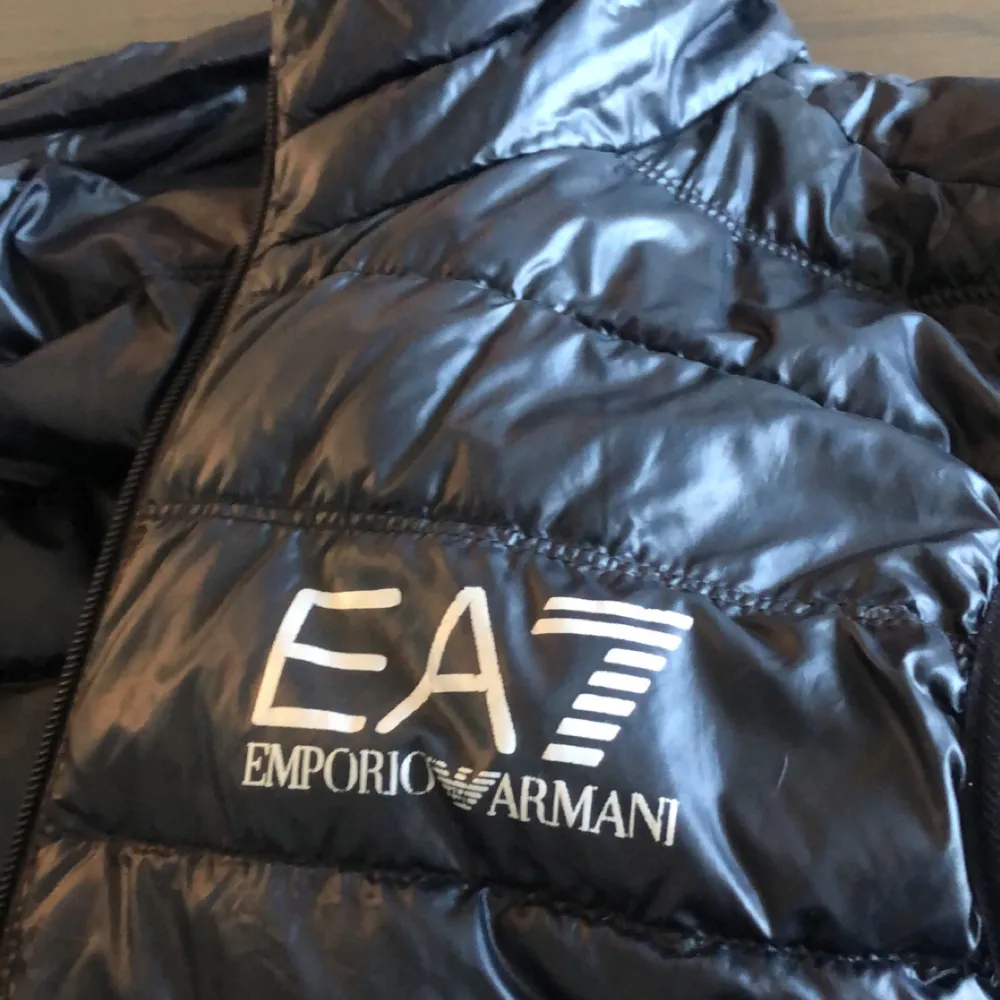 Säljer en snygg svart dunväst från Emporio Armani EA7. Västen har en glansig finish och är perfekt för kyligare dagar. Den har en dragkedja framtill och är quiltad för extra värme. Passar både till vardags och lite mer uppklädda tillfällen.. Jackor.