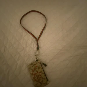 Svart plånbok med rem - Säljer en snygg svart plånbok med en cool rem i rött och grönt. Plånboken har en dragkedja och flera fack för kort. Perfekt för att hålla ordning på dina grejer när du är på språng!