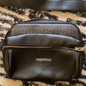 Svart axelväska från Valentino - Snygg svart axelväska från Valentino i syntetmaterial. Väskan har en justerbar rem och en praktisk dragkedja framtill. Perfekt för både vardag och fest! Den har också ett subtilt mönster med Valentino-loggan som ger en lyxig touch. Pris går och diskutera 😀