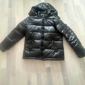 Svart pufferjacka med huva - Säljer en svart pufferjacka i mycket bra skick. Den är perfekt för kalla dagar med sin tjocka vaddering och har en praktisk huva. Jackan har dragkedja och två fickor med dragkedjor. Den är långärmad och passar bra för både höst och vinter.