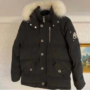 Svart dunjacka från Moose Knuckles - Säljer en svart dunjacka från Moose Knuckles i nyskick. Bara använder ett fåtal gånger. Jackan har en fluffig vit päls på huvan och flera praktiska fickor med knappar. Den är perfekt för kalla vinterdagar och har en snygg, stilren design med dragkedja och knappar framtill.