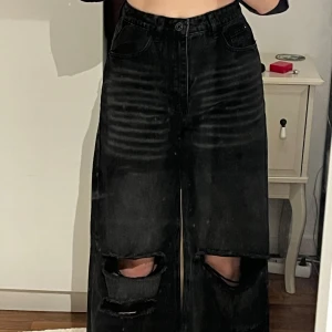Svarta jeans med slitningar - Säljer ett par svarta jeans i loose fit med coola slitningar på knäna. De är högmidjade och har en avslappnad, bred passform som är perfekt för en casual look. Materialet är denim och de har klassiska fem fickor och knappgylf. Perfekta för höst och vår!