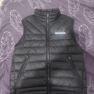 Svart dunväst från Emporio Armani - Säljer en snygg svart dunväst från Emporio Armani. Västen har en dragkedja framtill och är quiltad för extra värme. Perfekt för höst och vår när du vill hålla stilen utan att frysa. Den har en normal passform och är i mycket bra skick. Den är storlek S men passar även storlek M