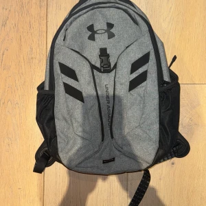 Grå ryggsäck från Under Armour - Säljer en snygg och praktisk grå ryggsäck från Under Armour, små tecken på den är använd (syns på bild nr5). Perfekt för både skola och träning. Den har flera fack, inklusive ett vadderat fack för laptop och sidofickor för vattenflaska. Justerbara remmar för bästa passform. Använd men i mycket bra skick! * Rymmer ca 31,5 liter  * Mått: 22,6 x 51 x 32 cm, * Rymmer upp till 15- och MacBook Pro eller liknande storlek,  * Vattenresistens, 100% polyester