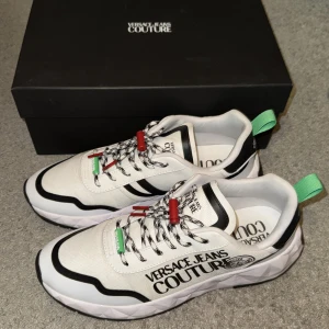 Versace Jeans Couture Sneakers - Snygga sneakers från Versace Jeans Couture i vitt och svart med coola detaljer i rött och grönt. Skorna har en modern design med snörning och är tillverkade i nylon och gummi. Perfekta för att ge din outfit en lyxig touch!