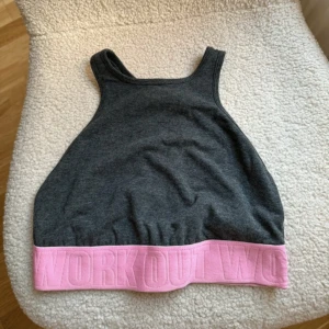 Grå och rosa sporttopp - Säljer en snygg grå sporttopp med rosa resårkant där det står 'WORK OUT'. Perfekt för träning. Toppen är ärmlös och har en tight passform. Superbekväm och stilren design!