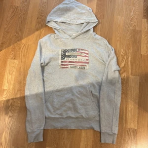 Grå hoodie från Ralph Lauren - Säljer en snygg grå hoodie från Ralph Lauren, Denim & Supply. Den har en cool patch med amerikansk flagga och text på framsidan. Bra skick, men har ett litet hål vid fickan, se bild.