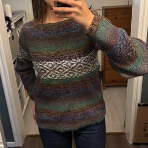 Flerfärgad stickad tröja - Säljer en supermysig flerfärgad stickad tröja med ett unikt mönster i vitt över bröstet. Tröjan har långa ärmar och en avslappnad passform, perfekt för höst och vinter. Den är i nyskick och redo att värma dig under kyliga dagar!