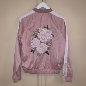 Bomberjacka med backprint/broderi - Suuperfin ljusrosa bomberjacka köpt på Ginatricot som snabbt blev slutsåld. Silvrig dragkedja och fickor med tryckknappar. Vita ränder längs ärmarna. Broderat blomtryck på ryggen. Storlek XS. Bra skick. Skriv vid intresse✨