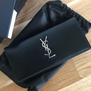 Plånbok, YVES SAINT LAURENT  - Svart plånbok i äkta läder (100% kalvskinn) med silvrigt monogram från YSL. Den är aldrig använd utan är i nyskick. Förpackning, tygpåse, äkthetsbevis och prislapp följer med. Nypris: 5 495kr 