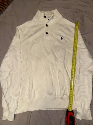 Vit stickad tröja från Ralph Lauren - Klassisk vit stickad tröja från Ralph Lauren med hög krage och tre bruna knappar. Tröjan har ribbade muddar och nederkant samt den ikoniska mörkblå polospelaren broderad på bröstet. Perfekt för en clean och stilren look.
