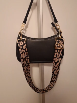 Svart axelväska med leopardband - Snygg svart axelväska i slätt konstläder med guldfärgade metalldetaljer. Väskan har ett brett axelband med leopardmönster i beige och svart, vilket ger en cool kontrast. Perfekt för dig som vill ha en trendig accessoar med attityd.