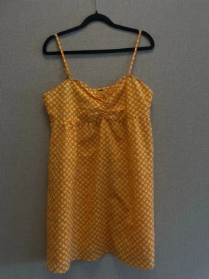 Gul blommig sommarklänning - Söt gul klänning med vita små blommor och tunna axelband. Klänningen har en lätt A-linjeform och är perfekt för varma dagar. Materialet känns lätt och luftigt, vilket gör den skön att bära under sommaren.