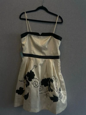 Champagnefärgad kort klänning med svarta detaljer - Snygg kort klänning i champagnefärg med svarta axelband och kant, samt svart band i midjan. Klänningen har blommigt mönster och paljettdetaljer nertill. Tillverkad i glansigt material och har smala axelband.