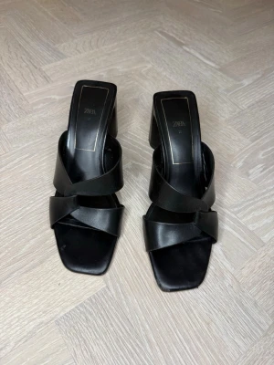 Svarta pumps med bred klack från Zara - Säljer ett par svarta pumps från Zara med bred klack och korsade remmar över foten. Skorna har fyrkantig tå och är tillverkade i slätt konstläder. Perfekta för dig som gillar stilrena och trendiga skor.