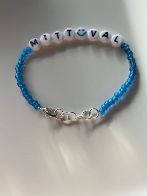 Blått pärlarmband med text - Ett handgjort armband med små blå pärlor och vita bokstavspärlor som stavar 'MITT VAL' med en smiley. Armbandet har en silverfärgad kedja och karbinlås för enkel justering. Perfekt för dig som vill ha ett personligt och färgglatt smycke.
