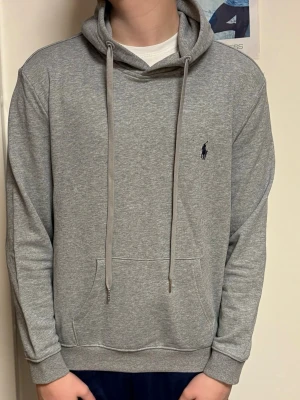 Grå hoodie från Polo Ralph Lauren - Snygg grå hoodie från Polo Ralph Lauren med klassisk huva och dragsnören. Broderad logga på bröstet och stor magficka framtill. Tillverkad i mjuk bomullsblandning som känns skön mot huden. Perfekt för en avslappnad och stilren look.