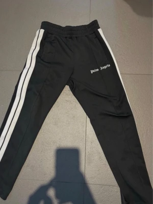 Palm Angels svarta mjukisbyxor - Palm Angels svarta mjukisbyxor med vita sidoränder och logga på vänster sida. Byxorna har resår i midjan och är tillverkade i ett glansigt, sportigt material. Perfekta för en avslappnad och trendig streetwear-look. Storlek s äkta såklart. Fast pris.