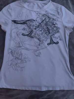 Vit t-shirt med svart blommönster - Säljer en vit t-shirt med ett stort, detaljerat svart blommönster och grafiskt tryck på framsidan. T-shirten har rund halsringning och korta ärmar. Materialet känns mjukt och bekvämt, perfekt för en avslappnad stil. Två små fläckar, ena på framsidan och andra på baksidan. Ingen lapp men troligtvis S eller XS