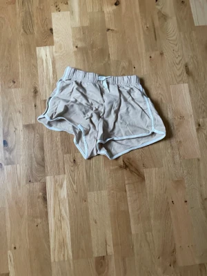 Beige shorts från H&M med vit kant - Säljer ett par beige shorts från H&M med vit kant och elastisk midja. De har en sportig look och är perfekta för varma dagar. Materialet känns mjukt och bekvämt, och passformen är loose för en avslappnad stil.