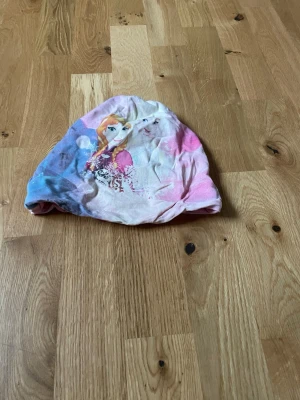 Färgglad Frozen mössa från Disney - Säljer en mjuk och färgglad mössa med motiv från Frozen och Anna på framsidan. Mössan har rosa, blå och vita toner och är tillverkad i ett stretchigt material som sitter skönt på huvudet. Perfekt för dig som älskar Disney och vill ha en unik accessoar.