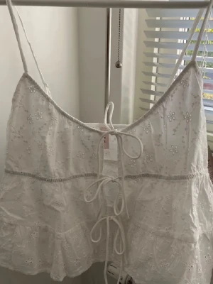 Vit broderad linnetopp med snörning - Söt vit linnetopp med tunna axelband och broderade blommor. Toppen har snörning i ryggen och volangdetaljer nedtill. Perfekt för varma dagar och ger en romantisk vibe till din outfit.