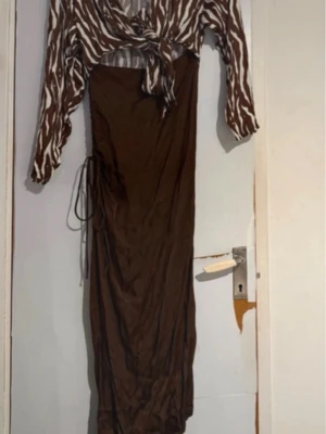 Brun långklänning med zebramönster - Säljer en brun långklänning med zebramönstrad överdel i vitt och brunt. Klänningen har långärmade ärmar och knyt framtill, samt snörningsdetalj i sidan. Perfekt för dig som vill sticka ut med en unik look.