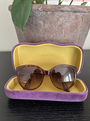 Bruna solglasögon från Gucci - Snygga solglasögon från Gucci med brunskiftande bågar i klassisk cat-eye form. Bågarna har ett diskret guldfärgat GG-logo på sidan och glasen är tonade i brunt. Kommer i en lyxig lila och gul sammetsfodral med Gucci-logga.  Modell: GG0631S Färgkod: 002 Storlek: 56-18 Skalmängd: 145  Knappt använda. 