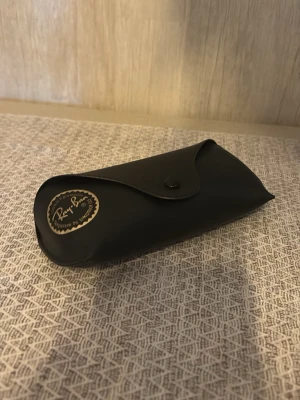 Ray-Ban svart fodral till solglasögon - Snyggt svart fodral från Ray-Ban till solglasögon, tillverkat i ett slätt material med guldfärgad logga och knappstängning. Perfekt för att skydda dina shades och hålla stilen. Har du tappat bort fodralet? Perfekt finns ett här nu!! (Dessa är från ett par rayban boyfriends ja nu tyvärr tappat bort) 