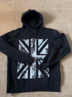 Svart hoodie med Union Jack-tryck - Snygg svart hoodie med stor Union Jack-flagga i grått och vitt på framsidan. Hoodien har huva med dragsko och långa ärmar. Perfekt för dig som gillar streetwear och vill sticka ut med en cool grafisk design.