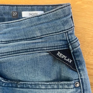 Blå raka jeans från Replay - Säljer ett par klassiska blå jeans från Replay i modellen Jacksy. Jeansen har raka ben, fem fickor och snygga slitningar för en avslappnad look. Tillverkade i mjuk denim med tydliga sömmar och Replay-logga på myntfickan.