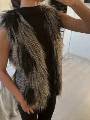 Svartgrå lång pälsväst - Säljer en snygg svartgrå pälsväst med längre, fluffig fuskpäls och öppen front. Västen har en cool, mönstrad look med inslag av ljusare och mörkare toner som ger en trendig vibe. Perfekt att slänga över en enkel outfit för att höja stilen.