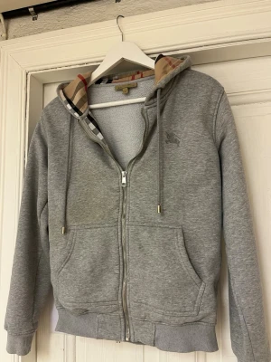 Burberry hoodie - Snygg grå hoodie från Burberry med klassiskt rutigt foder i huvan och dragkedja framtill. Hoodien har två fickor, ribbade muddar och justerbar huva med snören. Perfekt för en stilren och avslappnad look.