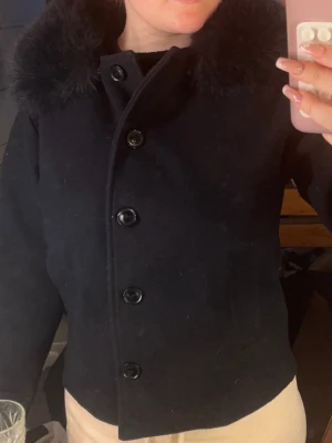 Pälsjacka från Gina Tricot  - Snygg svart pälsjacka med stor fluffig fuskpälskrage och stora svarta knappar framtill. Jackan har rak passform och långa ärmar med knappdetalj vid ärmslut. Perfekt för kalla vinterdagar och ger en stilren look.