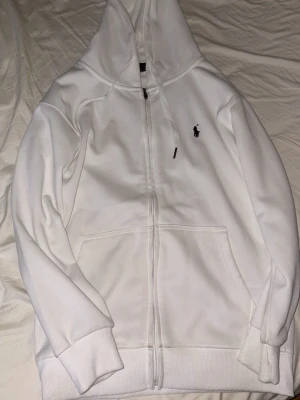 Vit hoodie från Polo Ralph Lauren - Säljer en clean vit hoodie från Polo Ralph Lauren med dragkedja, huva och kängurufickor. Klassisk logga broderad på bröstet. Tillverkad i mjukt bomullsmaterial som känns skönt mot huden. Perfekt för en avslappnad och stilren look.