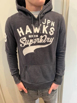 Mörkgrå Superdry hoodie med tryck - Snygg mörkgrå hoodie från Superdry i storlek M modellen är 173 cm och väger 62kg