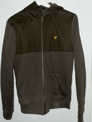 Olivgrön hoodie från Lyle & Scott - Snygg olivgrön hoodie med dragkedja från Lyle & Scott. Har huva, känguruficka och den klassiska gula loggan på bröstet. Tillverkad i mjukt bomullsmaterial, perfekt för en avslappnad stil.