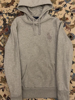 Grå hoodie från Polo Ralph Lauren - Snygg grå hoodie från Polo Ralph Lauren med stor broderad logga på bröstet. Klassisk modell med huva och dragsnören samt känguruficka framtill. Tillverkad i mjuk bomullsblandning som känns skön mot huden. Perfekt för en avslappnad och stilren look.