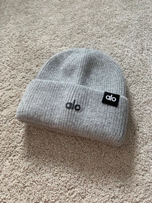 Alp Yoga Mohair/Alpacka Mössa - Snygg grå ribbstickad mössa från Alo med uppvikt kant och svart logotyp framtill. Tillverkad i mjukt och värmande material som passar perfekt för kyliga dagar. Klassisk och stilren design som funkar till det mesta.
