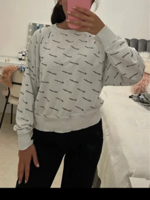 Vit sweatshirt från Champion - Säljer en vit sweatshirt från Champion med svart logotypmönster över hela tröjan. Tröjan har rund halsringning, långa ärmar och ribbade muddar vid ärmslut och nederkant. Perfekt för en avslappnad och sportig stil.