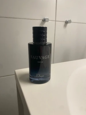Dior Sauvage Parfum 100ml - Säljer en elegant parfymflaska från Dior, modellen Sauvage Parfum. Flaskan är mörkblå med svart lock och har en stilren, cylindrisk form. Texten på flaskan är i vitt och ger ett lyxigt intryck. Perfekt för dig som gillar exklusiva dofter.