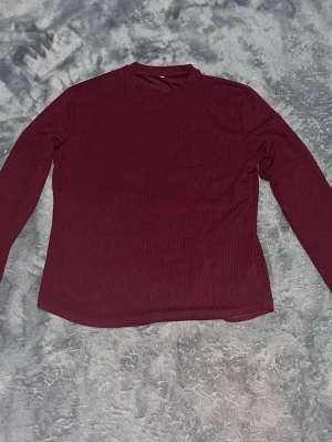 Vinröd ribbad långärmad tröja - En enkel och stilren vinröd långärmad tröja med ribbad struktur. Tröja har en rak passform och hög hals, Materialet känns mjukt och stretchigt för extra komfort.