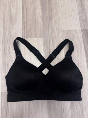Svart sport-bh med korsade band - Säljer en svart sport-bh med vadderade kupor och bred resår under bysten. Bh:n har justerbara axelband som korsas i ryggen för extra stöd. Materialet är stretchigt och har små ventilerande hål för bättre komfort.