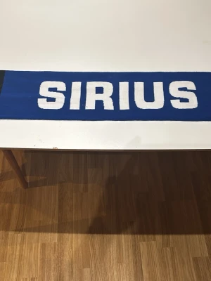 Blå halsduk med SIRIUS-tryck - Snygg blå halsduk med stort vitt SIRIUS-tryck. Perfekt accessoar för dig som vill visa ditt stöd för Sirius. Halsduken är rektangulär och har en klassisk supporterstil. Materialet känns mjukt och bekvämt mot huden.