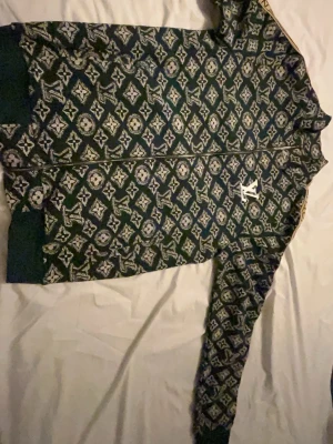 Mönstrad ziptröja från Louis Vuitton - Säljer en grön ziptröja från Louis Vuitton med ikoniskt monogrammönster i vitt och blått. Tröjan har hel dragkedja, ribbade muddar och ståkrage. Perfekt för dig som gillar streetwear och exklusiva detaljer.