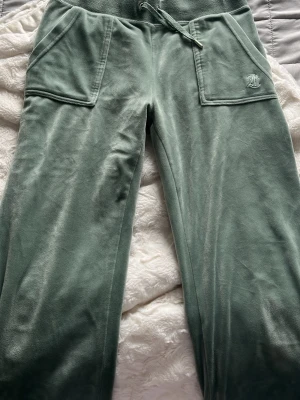 Gröna velourbyxor Juicy Couture - Mjukisbyxor i grön velour från Juicy Couture med breda raka ben, stora fickor fram och bak samt dragsko i midjan. Ikonisk JC-logga broderad på ena fickan framtill. Perfekta för en chill och trendig look.